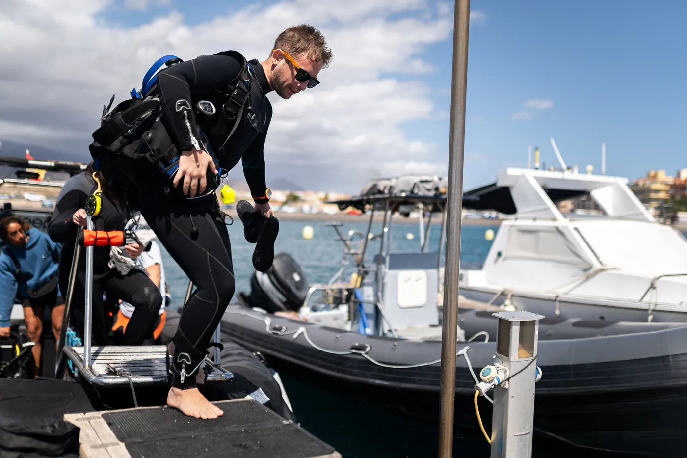 A diver in full gear stepping from the pier onto a dive boat, with other divers getting ready behind him